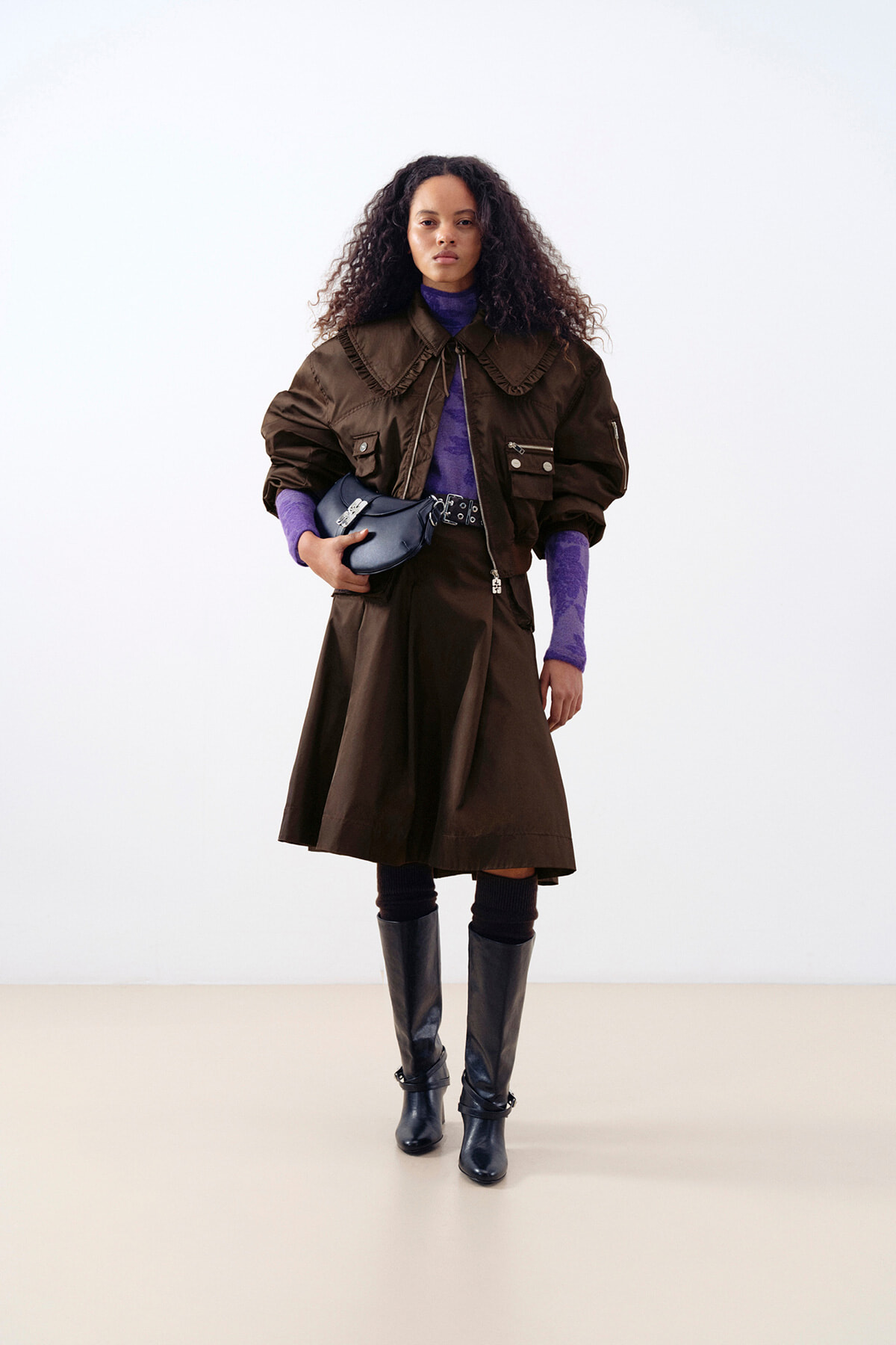 Woman with curly hair wearing a brown oversized coat, purple top, and black knee-high boots, holding a blue clutch while standing against a light background.