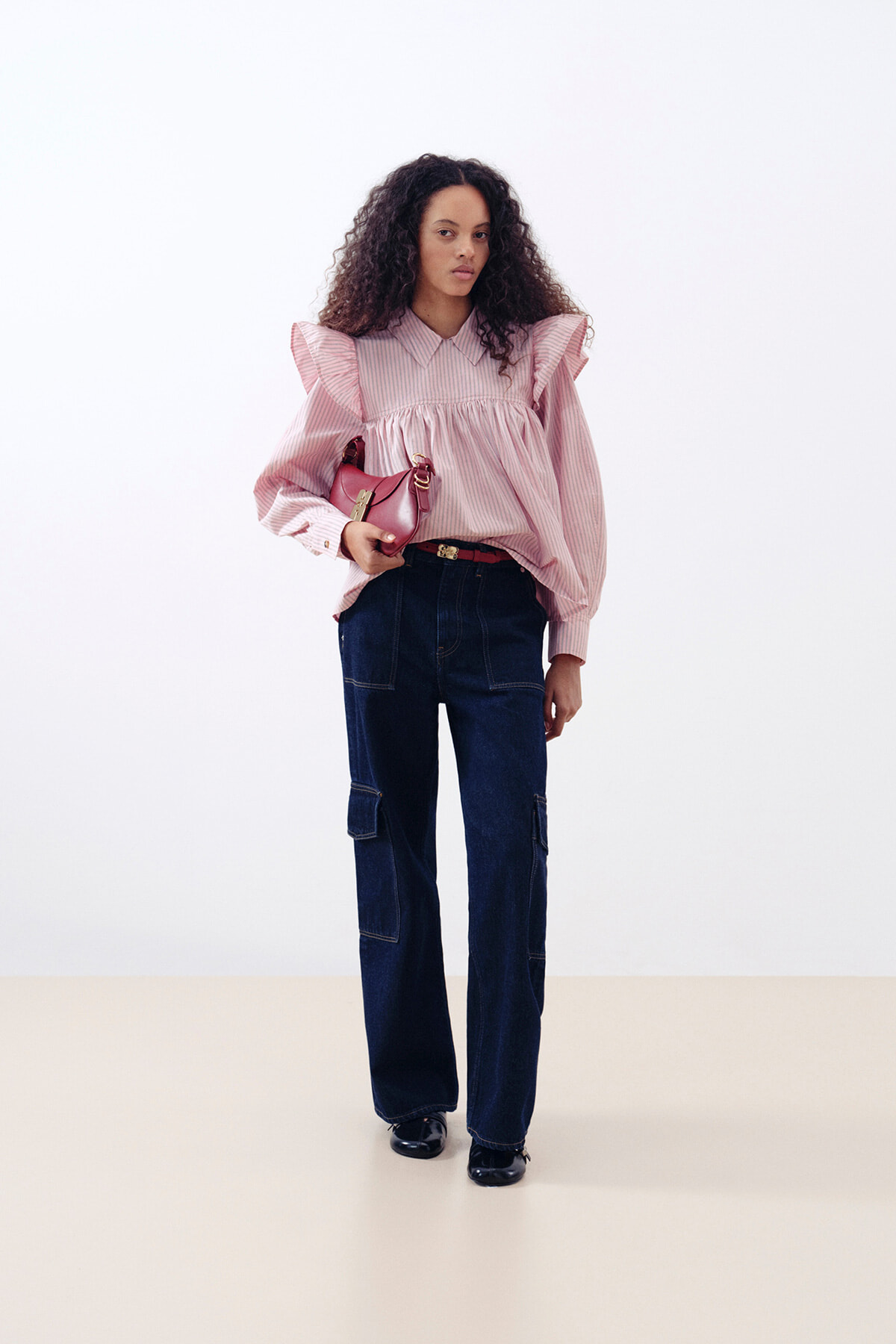 Woman standing in a pink striped blouse with ruffled shoulders, dark wide-leg jeans, and black shoes, holding a small pink purse.