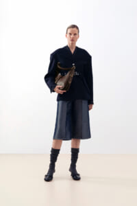 Woman in a navy blazer with belt, holding a brown shoulder bag, standing against a light background; mid-length denim skirt and gray leg warmers with black shoes.