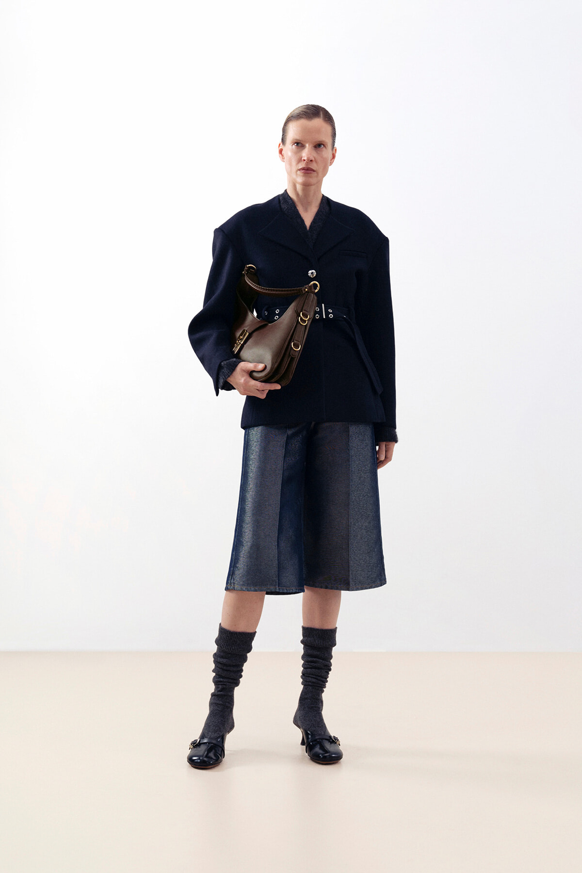 Woman in a navy blazer with belt, holding a brown shoulder bag, standing against a light background; mid-length denim skirt and gray leg warmers with black shoes.