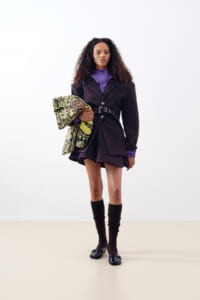 Model in a dark belted coat over a purple blouse, with a short skirt and brown knee-high socks, holding a patterned knit coat under her arm in a studio.