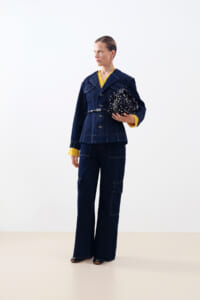 Full-body shot of a person in a dark denim suit with yellow cuffs, holding a black-and-white patterned bag.