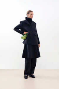 Person in a dark tailored blazer with a matching loose skirt over wide-leg pants, wearing a black turtleneck. They hold a small metallic green handbag.
