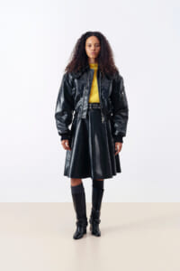 Model wearing a black leather jacket over a matching leather skirt with a yellow top peeking out, paired with knee-high boots in a studio setting.