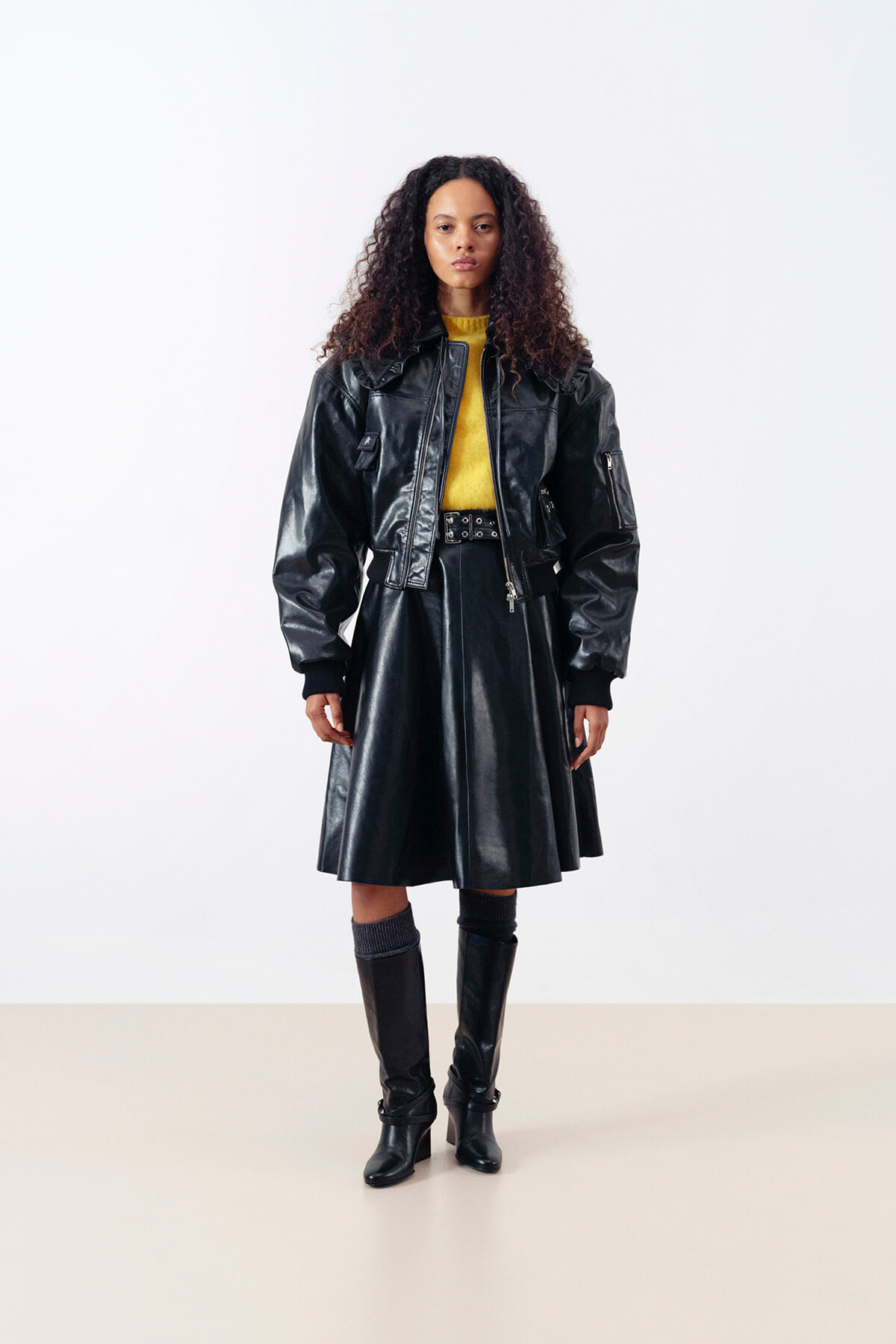 Model wearing a black leather jacket over a matching leather skirt with a yellow top peeking out, paired with knee-high boots in a studio setting.