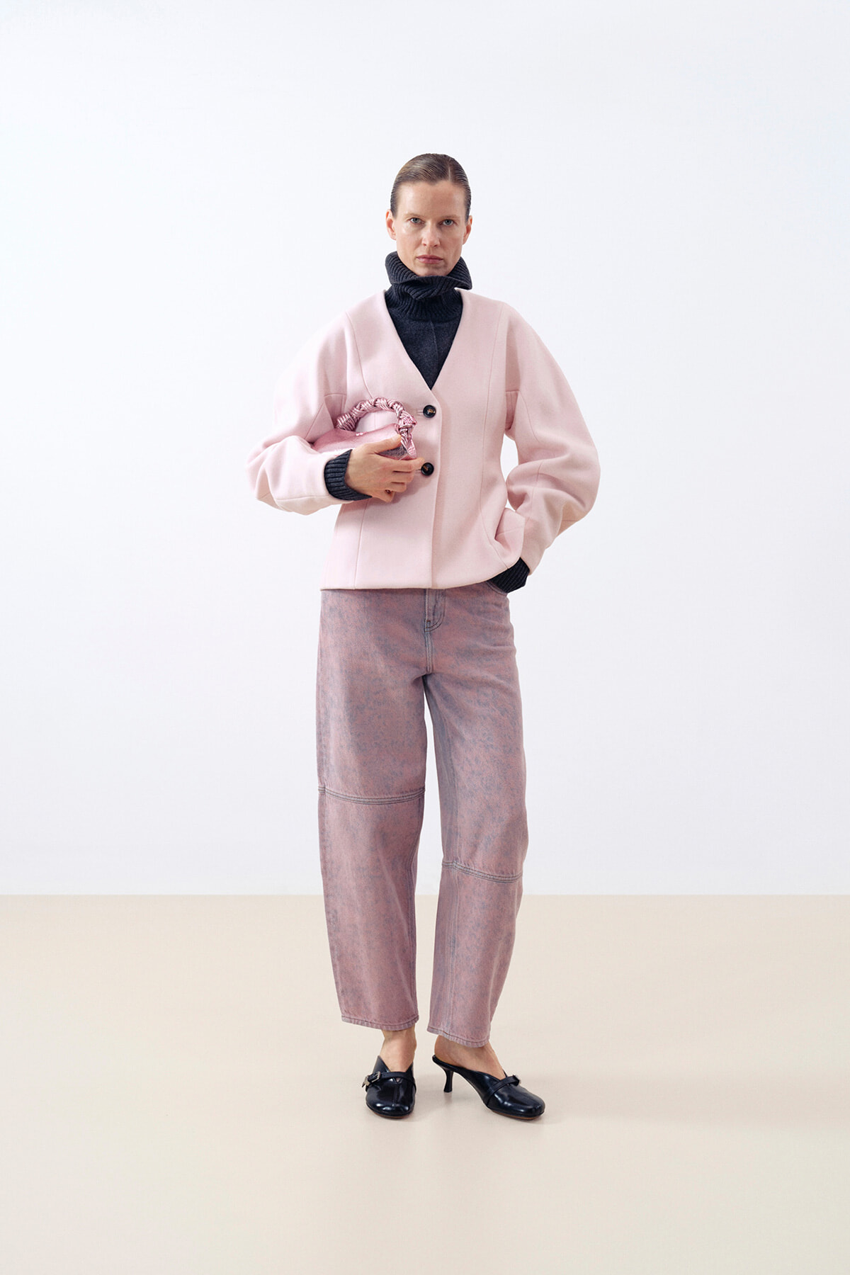 Model in a pale pink coat over a dark turtleneck, pink-washed jeans, and black heels, standing against a light backdrop and holding a pink braided accessory