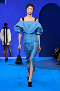 Model on a blue runway wearing a light blue off‑the‑shoulder dress with oversized puff sleeves and a front slit, carrying a black handbag.