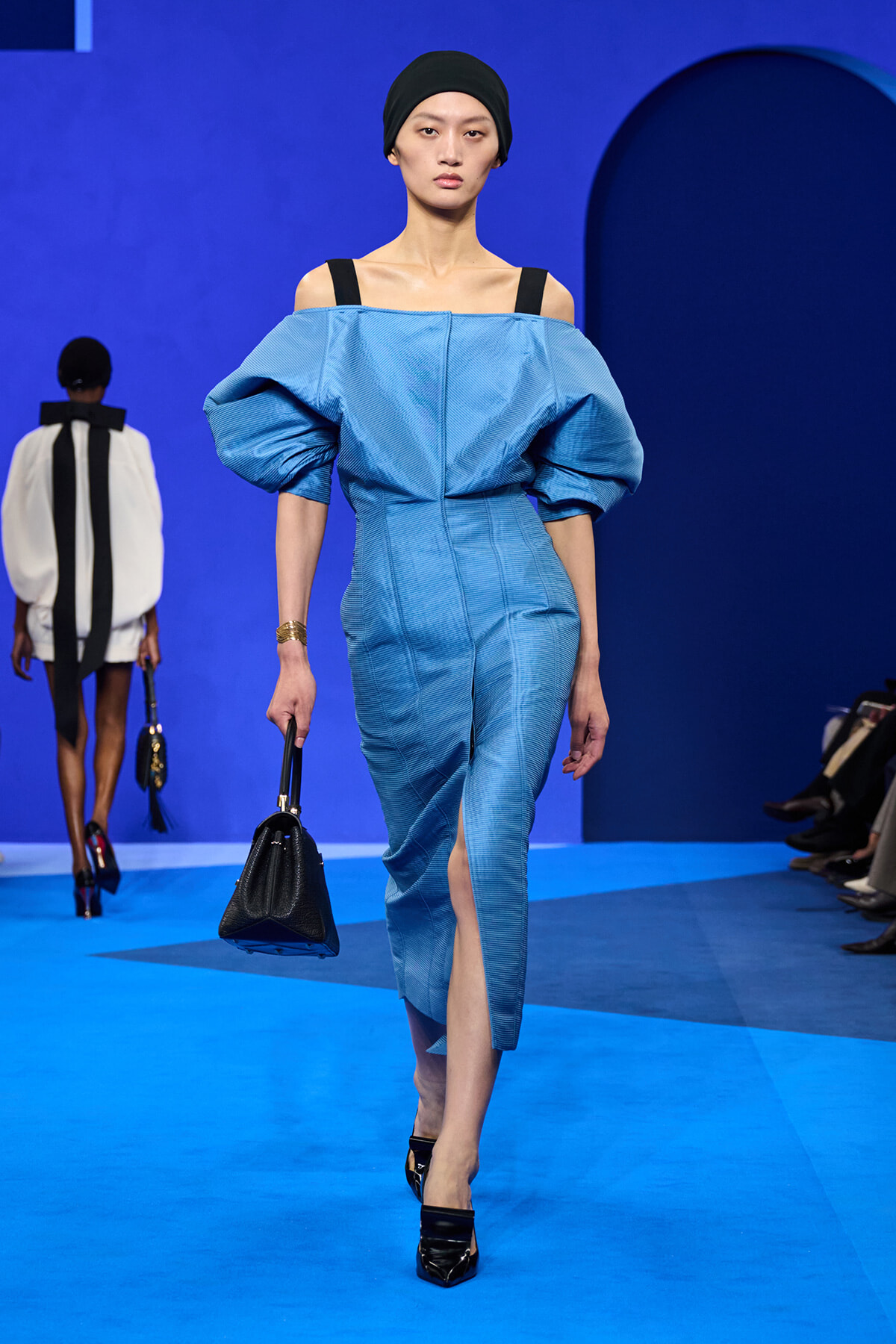 Model on a blue runway wearing a light blue off‑the‑shoulder dress with oversized puff sleeves and a front slit, carrying a black handbag.
