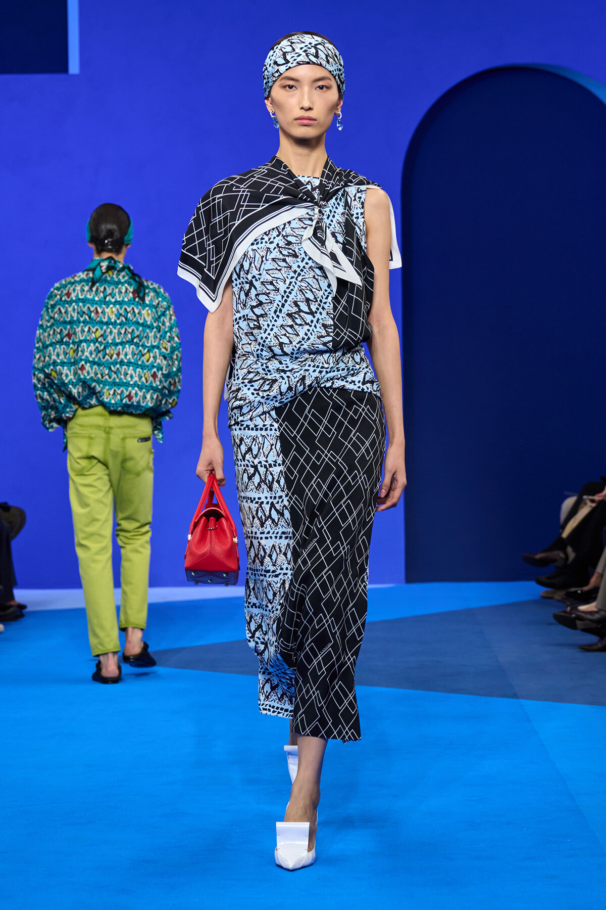 Model walking a runway in a blue-black patterned dress with a matching headscarf, holding a red handbag and wearing white heeled shoes.