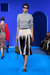 Model walks the runway in a gray turtleneck sweater and a black-and-white angular midi skirt, carrying a lime-green bag with tassel earrings.