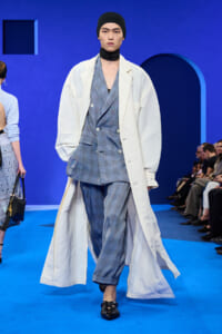 Male model walking a fashion runway in a blue plaid suit with an oversized white coat, black beanie, and neck scarf, striking pose.