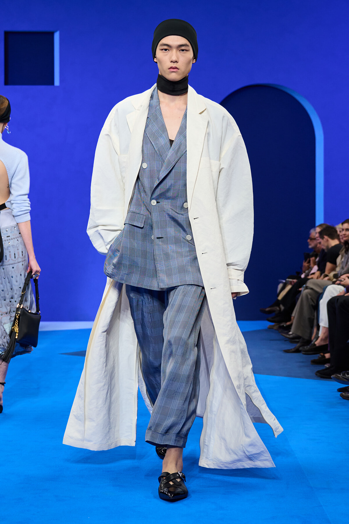Male model walking a fashion runway in a blue plaid suit with an oversized white coat, black beanie, and neck scarf, striking pose.