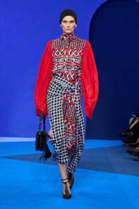 Model on a blue runway wearing a red, black, and white patterned dress with billowy red sleeves and a black headscarf, carrying a black handbag.