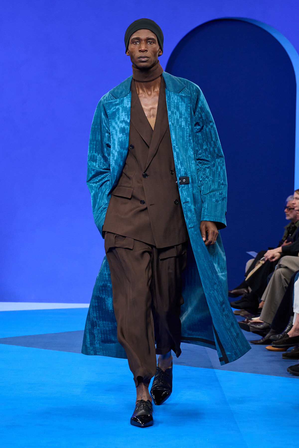 Male model walks a blue runway in a teal patterned long coat over a brown double-breasted suit, wearing a black beanie against a blue arch backdrop.