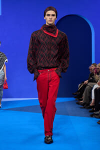 Male model on a blue runway wearing a black and red geometric-patterned top, red scarf, red pants, and matching headband; hands in pockets.