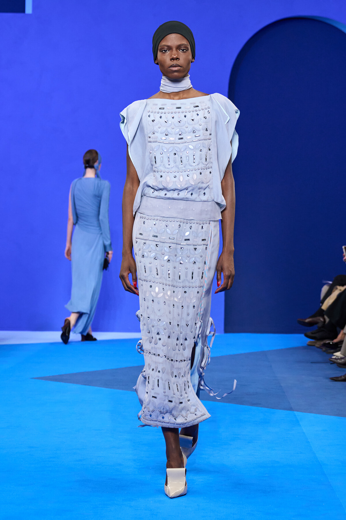 Model walks a blue runway wearing a light-blue embellished sleeveless dress with a matching neck scarf and black head wrap.