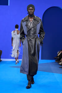 Model wearing a two-tone black and blue leather trench coat with a belt, black beanie, walking on a blue runway stage.