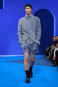 Male model walks a runway in a light blue checkered suit with matching shorts, black beanie, and black socks with loafers against a blue backdrop.