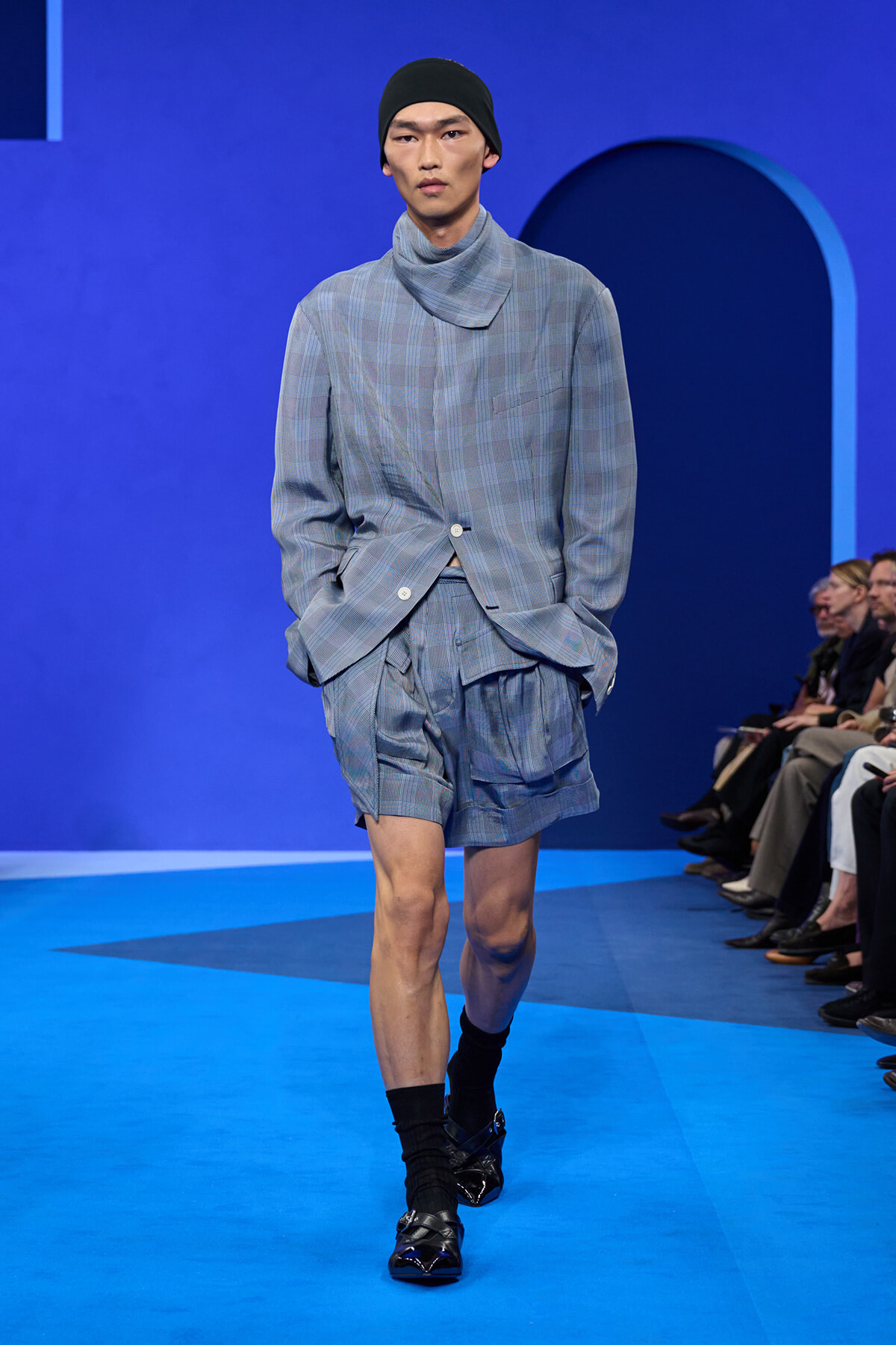 Male model walks a runway in a light blue checkered suit with matching shorts, black beanie, and black socks with loafers against a blue backdrop.