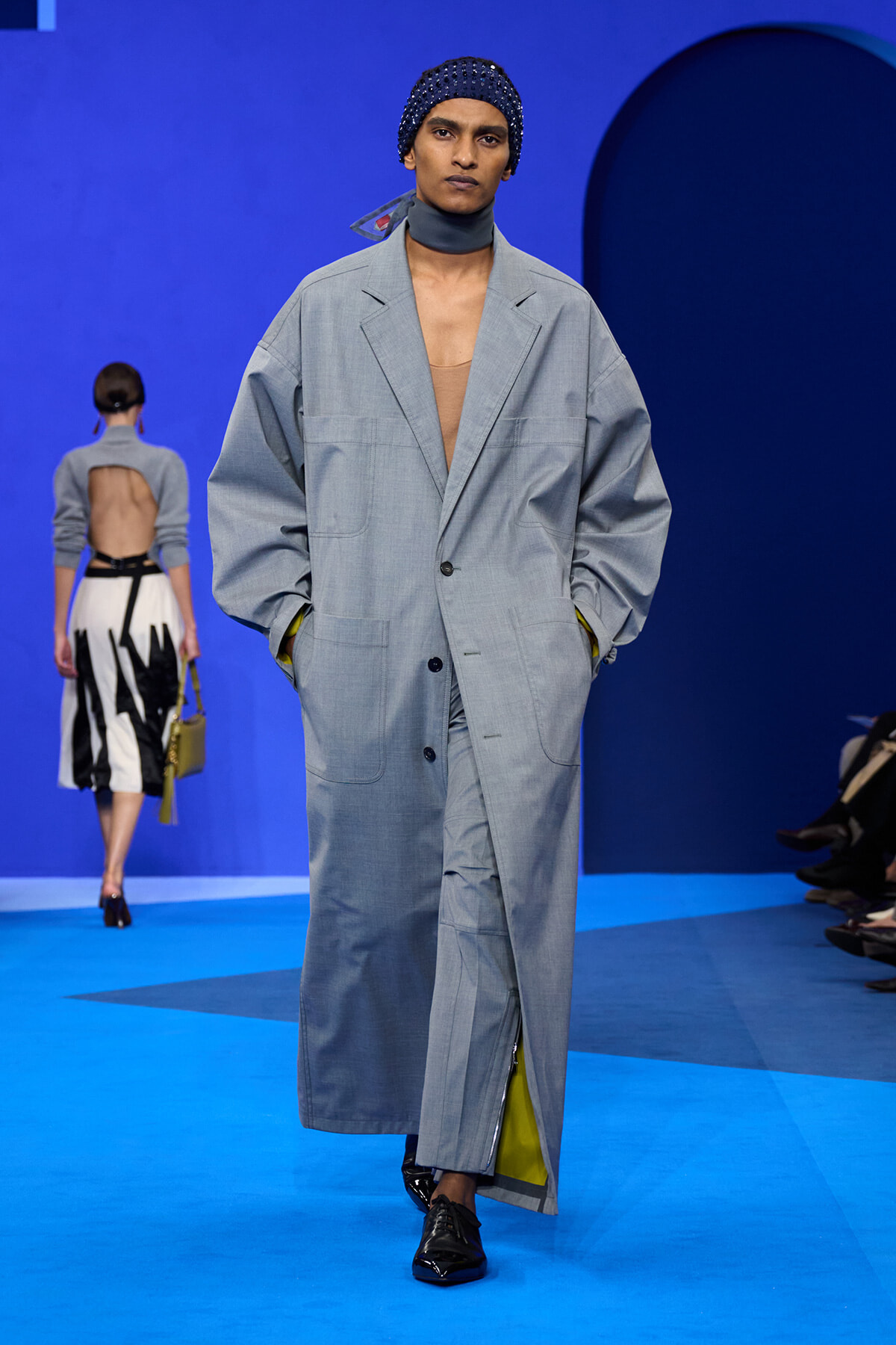 Model walks the runway in an oversized light-gray coat with a beige top, blue headscarf, and yellow lining peeking out, on a bright blue stage.