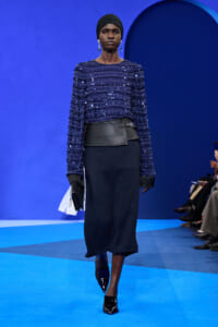 Model walks a blue-toned runway in a sequined navy top, wide black belt, and dark midi skirt with patent black heels and a clutch.