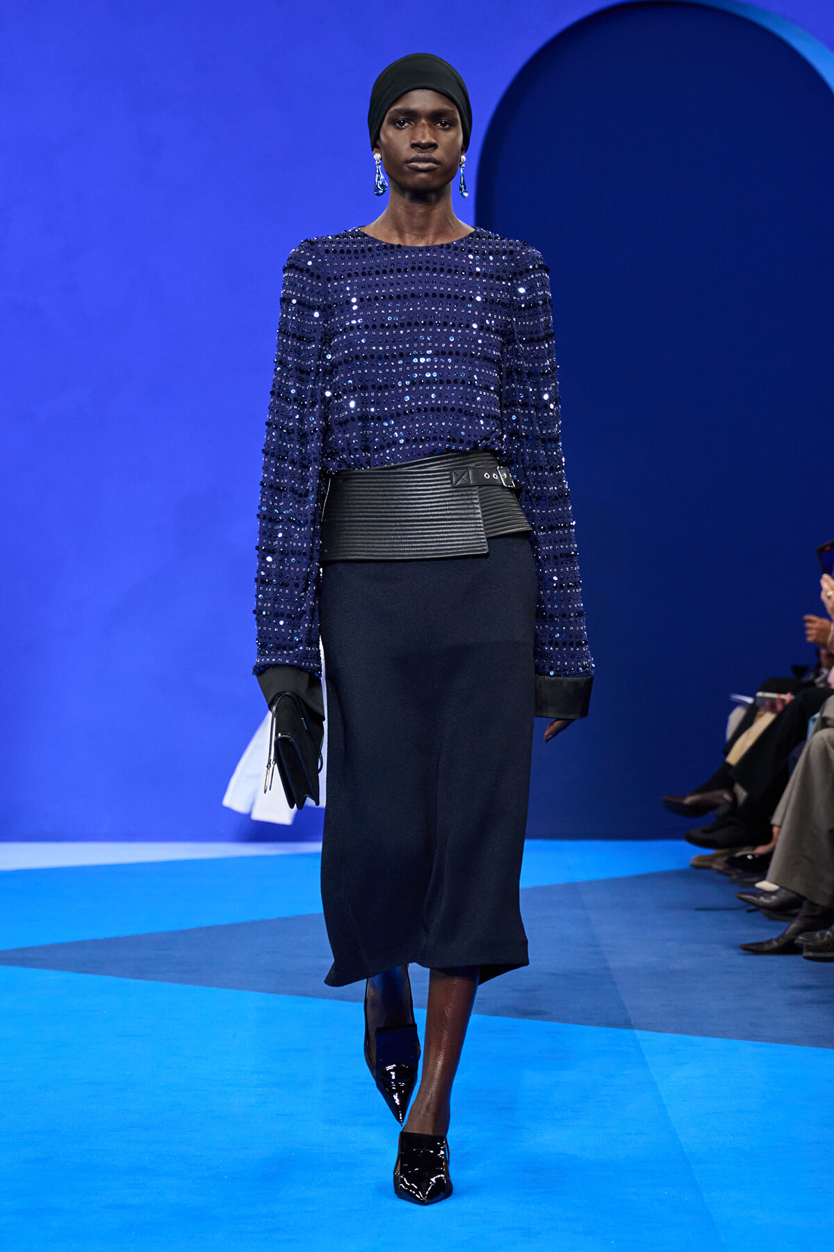 Model walks a blue-toned runway in a sequined navy top, wide black belt, and dark midi skirt with patent black heels and a clutch.