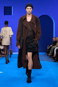 Model walking a fashion show in a long brown coat over a dark outfit, black beanie, and black shorts on a blue runway with a quilted clutch.