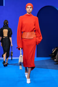 Model in a bright red midi dress with oversized sleeves, matching wide belt, red headscarf, and white handbag on a blue runway.