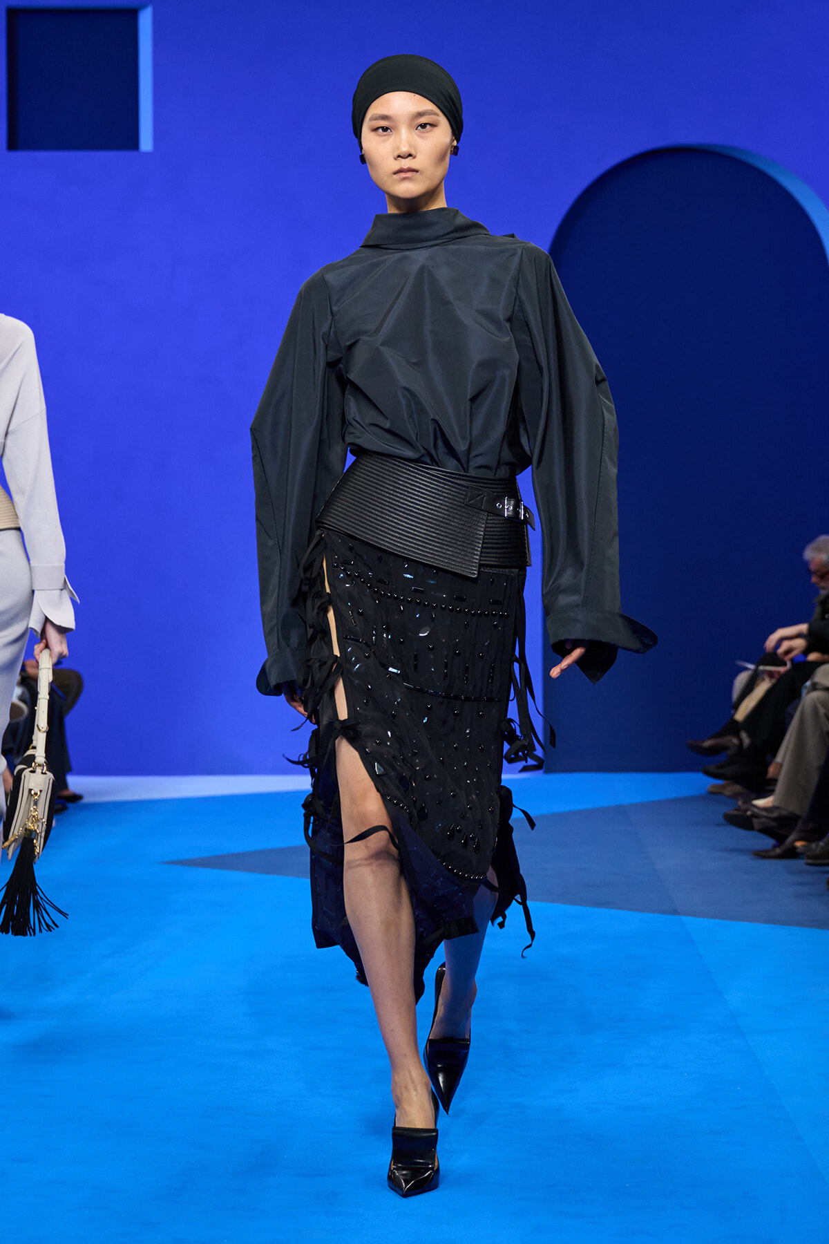 Model walking a fashion show in a dark, oversized blouse with a wide black belt and a black beaded skirt with side slits; black head wrap, black pointed heels, blue runway backdrop.