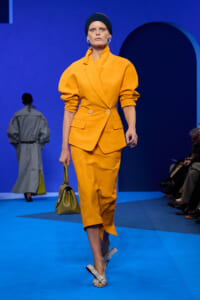 Model in a bright orange-yellow suit with a double-breasted blazer and matching skirt walks a blue runway, carrying an olive handbag.