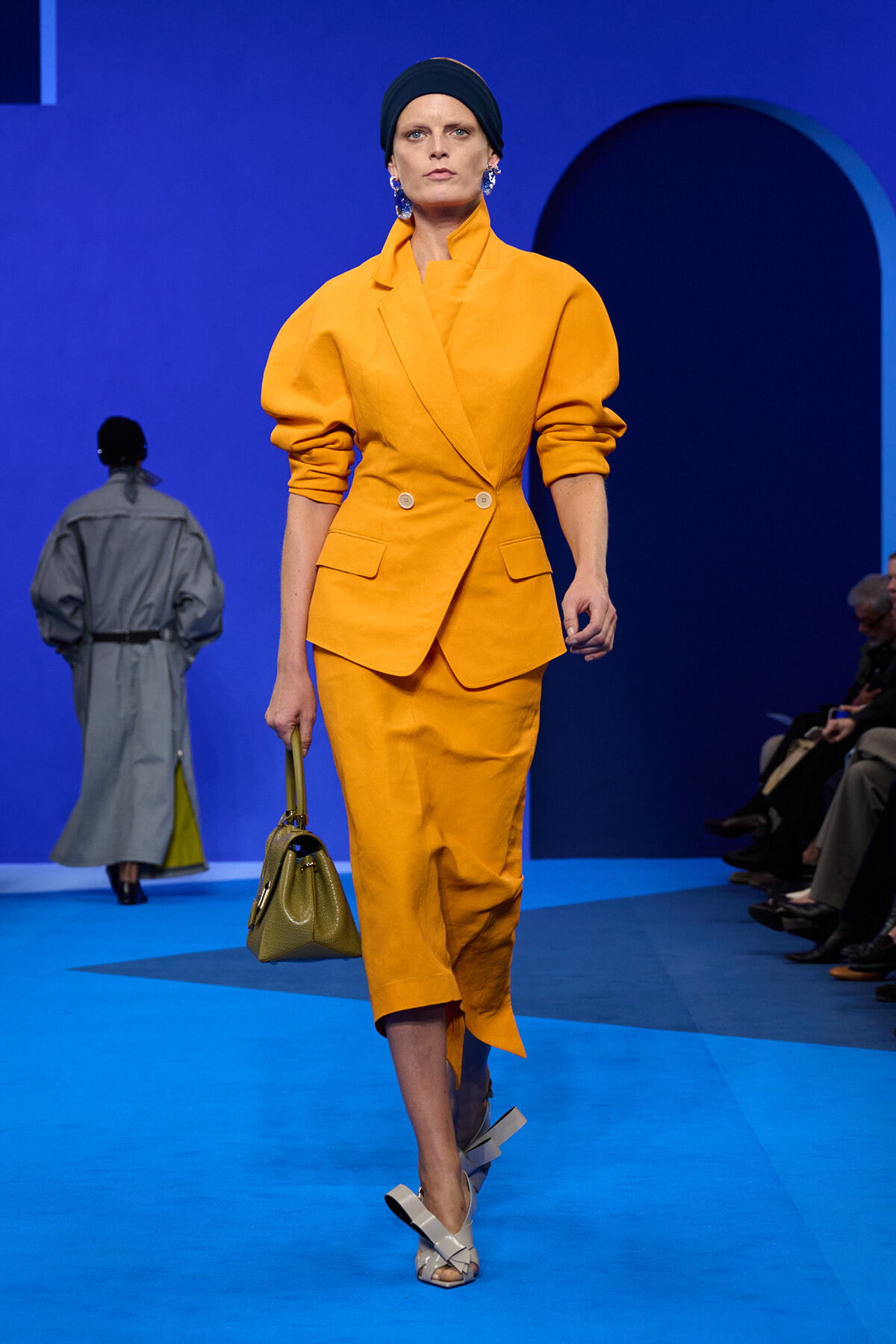Model in a bright orange-yellow suit with a double-breasted blazer and matching skirt walks a blue runway, carrying an olive handbag.