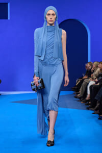 Model on a blue runway wearing a matching blue draped gown and hijab, carrying a geometric black clutch.