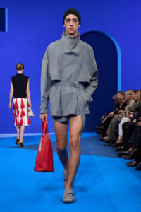 Male model walking a blue runway in a gray capeTop with belt, gray shorts, black beanie, and gray shoes; carrying a red handbag.