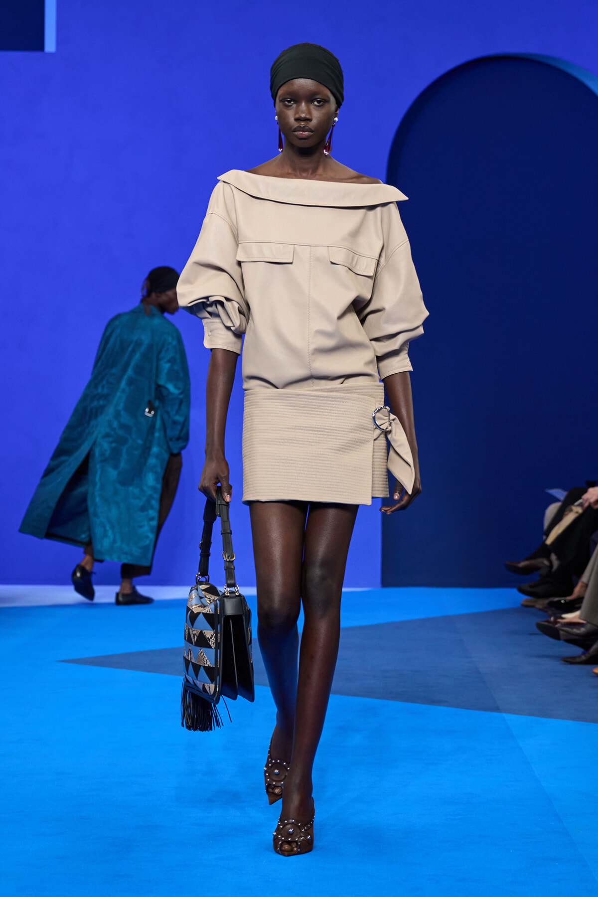 Model wearing a beige off-the-shoulder top with puff sleeves and a matching mini skirt, black headwrap, carrying a geometric patterned bag with fringe on a blue runway.