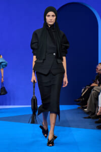 Model in a black tailored blazer and matching skirt walks a blue runway, wearing a hijab and carrying a black fringe handbag.