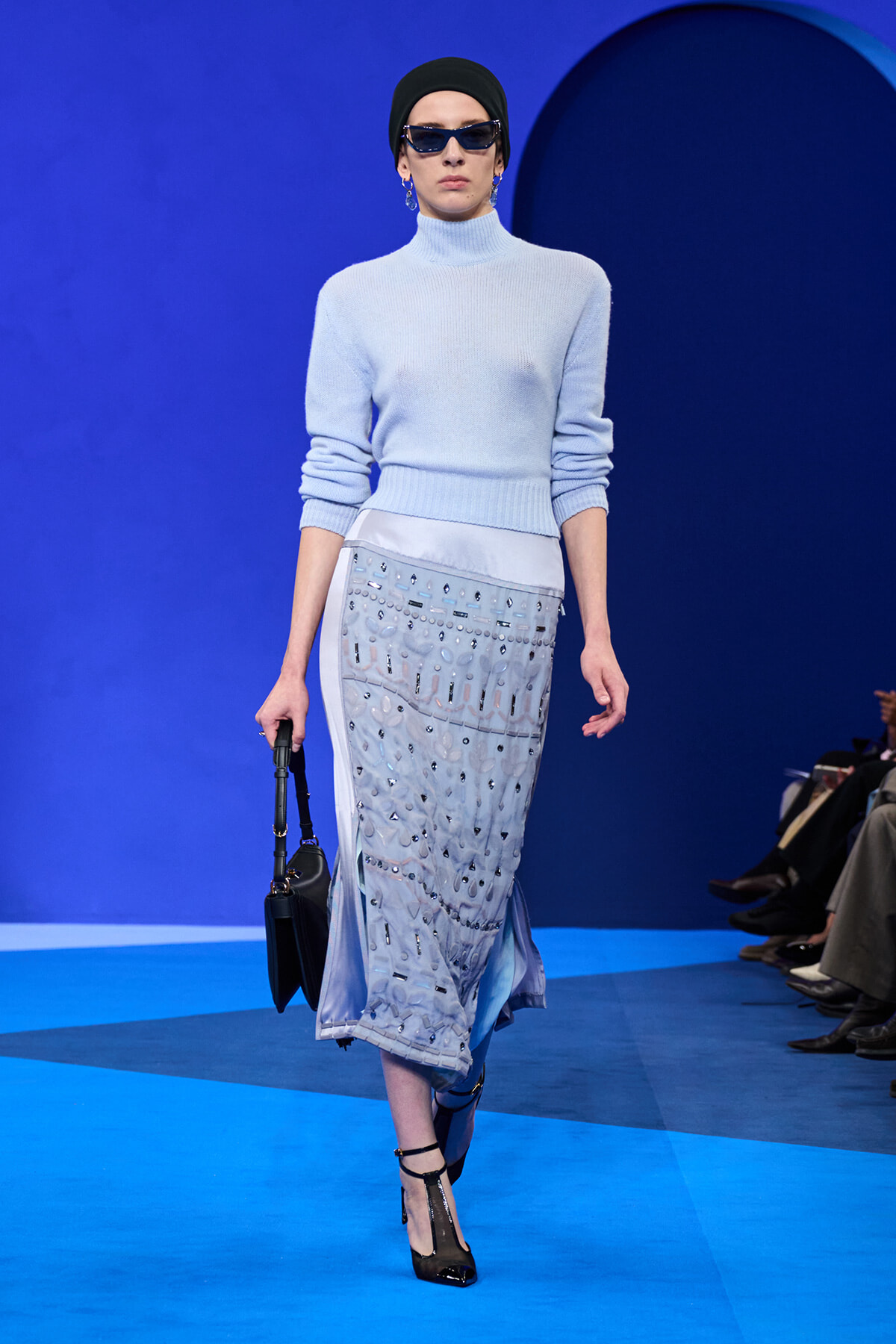 Model walks a blue runway in a pale blue knit turtleneck sweater and a silver-embellished midi skirt, with a black headwrap, sunglasses, and a small black handbag.
