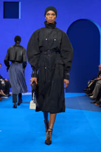 Model on a blue runway wearing a black belted trench-like coat and head wrap, carrying a small black bag with a tag in hand.