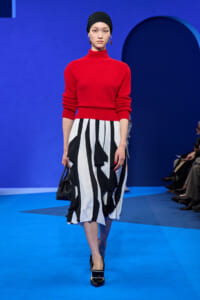 Runway model wearing a red knit turtleneck and a black-and-white striped, ruffled midi skirt, with a black headscarf and Heels, carrying a small black handbag on a blue catwalk.