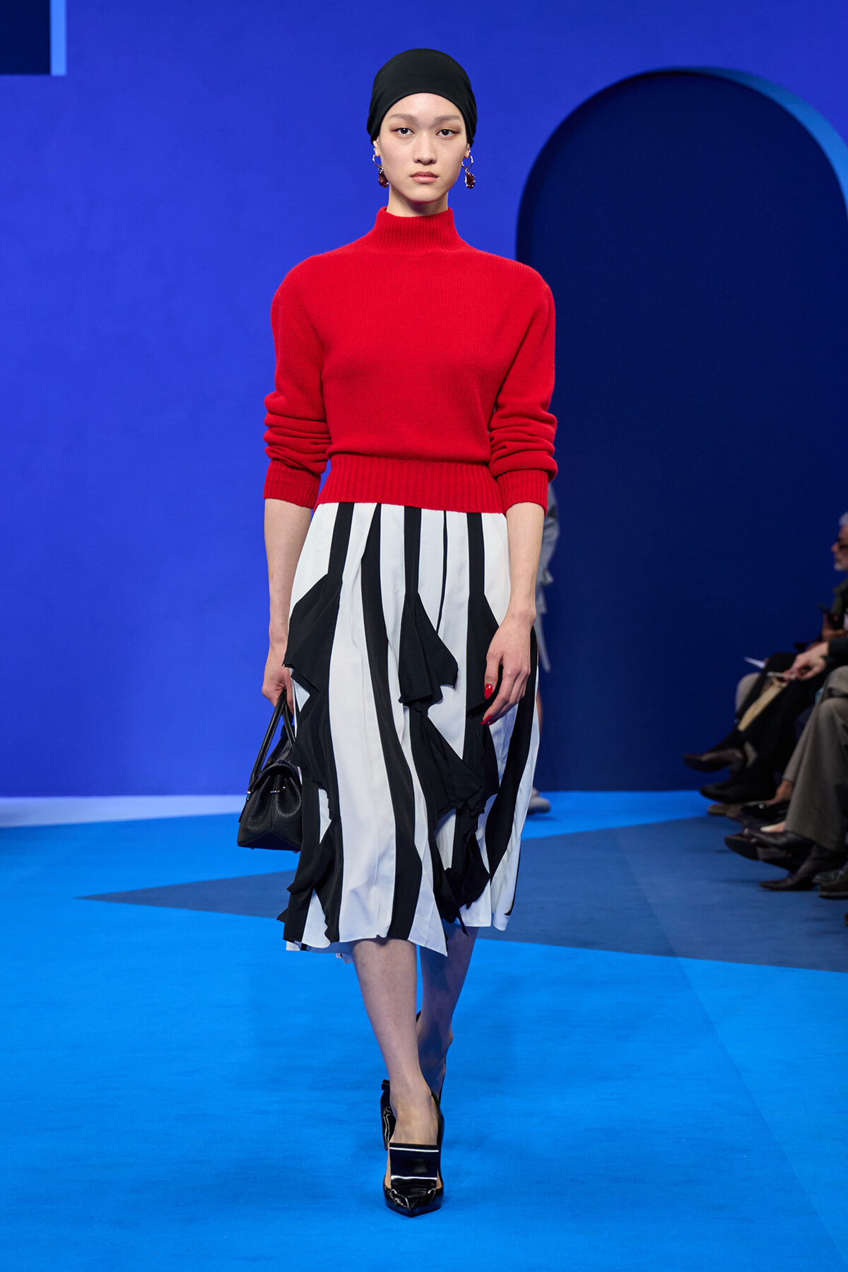 Runway model wearing a red knit turtleneck and a black-and-white striped, ruffled midi skirt, with a black headscarf and Heels, carrying a small black handbag on a blue catwalk.