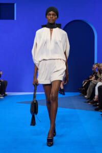 Model on a blue runway wearing an ivory draped blouse and matching shorts, with a black head wrap and a black woven handbag with tassel.