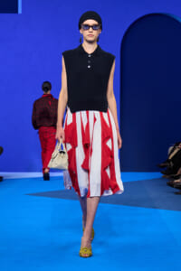 Runway model in a sleeveless black knit top, red-and-white striped midi skirt, yellow heels, sunglasses, and a beige handbag against a blue backdrop.