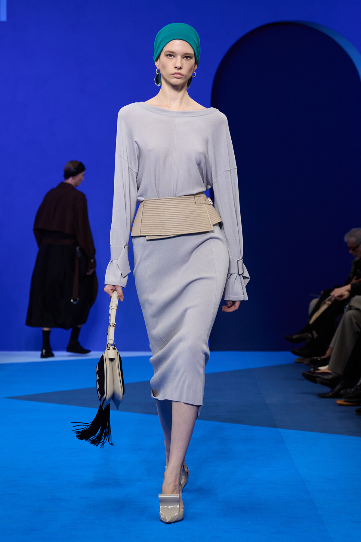 Model on a blue runway wearing a light gray knit dress with a beige waist belt, green headscarf, and a white handbag with a black fringe tassel