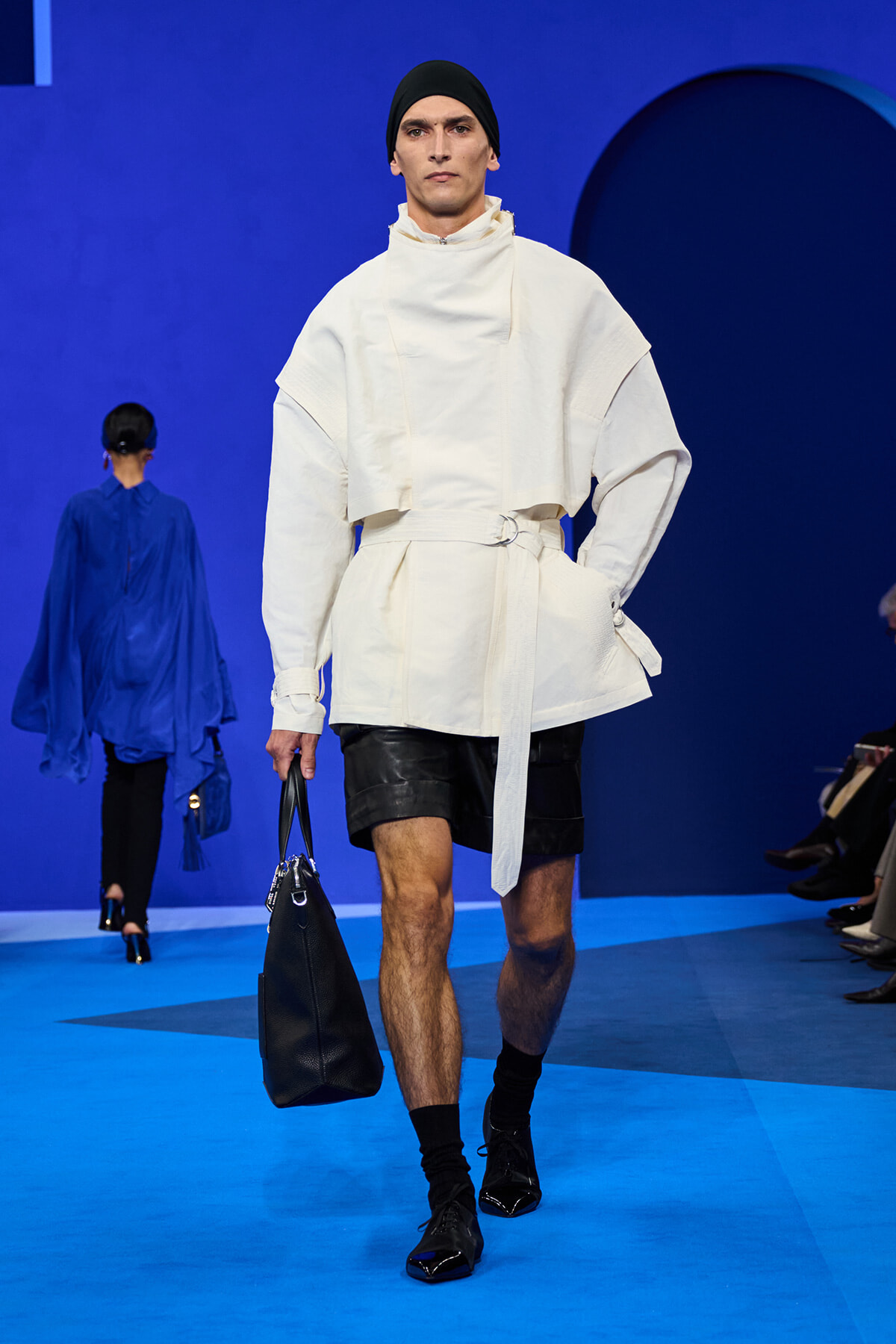 Male runway model in a white oversized top with belt, black shorts, and a black handbag on a blue catwalk.",