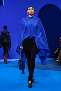 Model walks the runway in a cobalt blue oversized top, matching headscarf, and black pants, carrying a blue handbag with tassel.