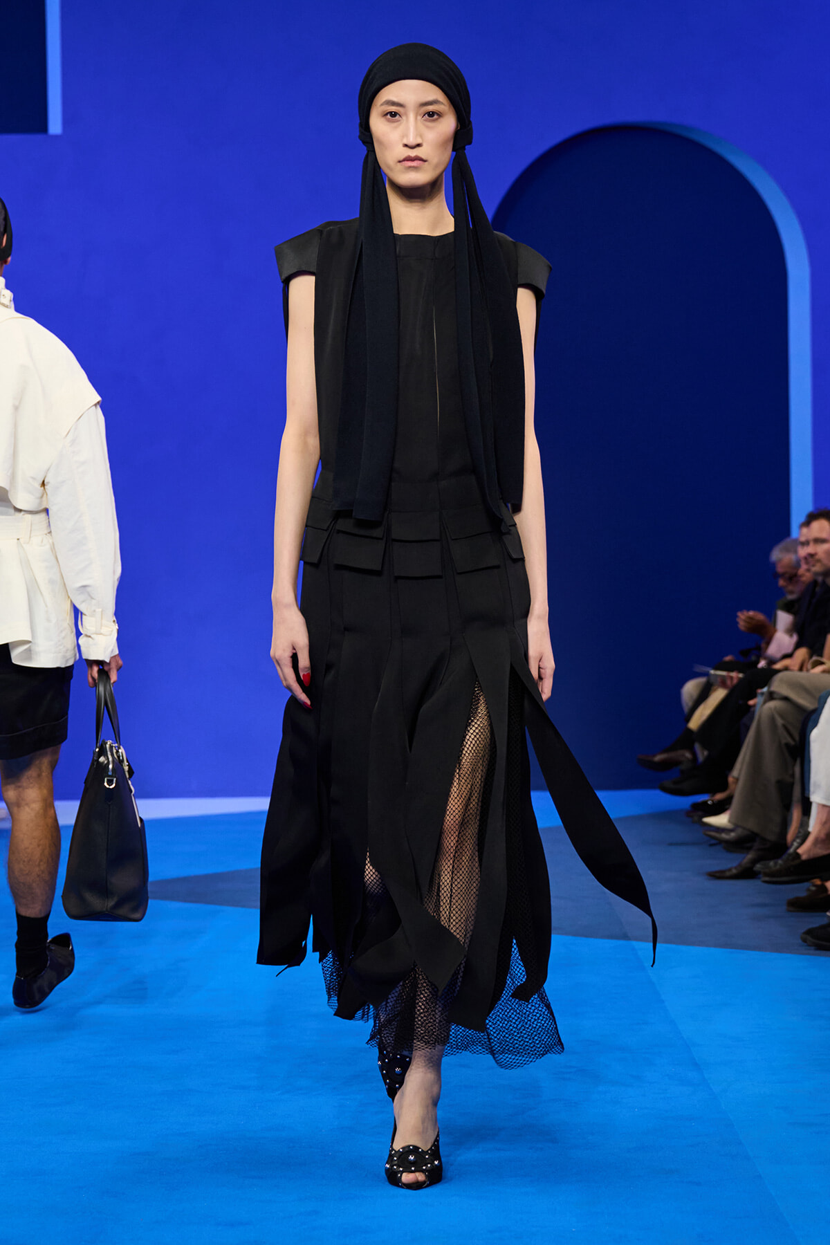 Model in an all-black outfit with a headscarf walking a blue runway, layered skirt and mesh underskirt visible near the hem.