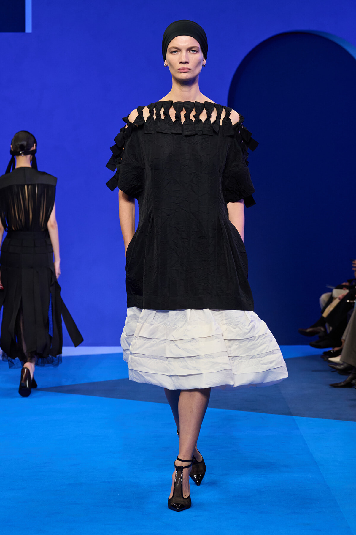 Model walking the runway in a black off-the-shoulder top with fringe cutouts and a white tiered skirt against a blue backdrop with heels.