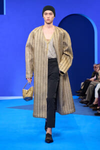 Male model walks a blue runway in a long metallic gold chevron coat over a beige top and black pants, wearing a black beanie and carrying a small gold handbag.
