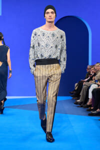 Male model walking a blue runway in a silver embellished geometric top and metallic gold trousers, with a black waistband and black beanie.