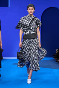 Model on a blue runway wearing a black-and-white patterned dress with a matching headscarf and bold belt, carrying a small studded bag and white heels.