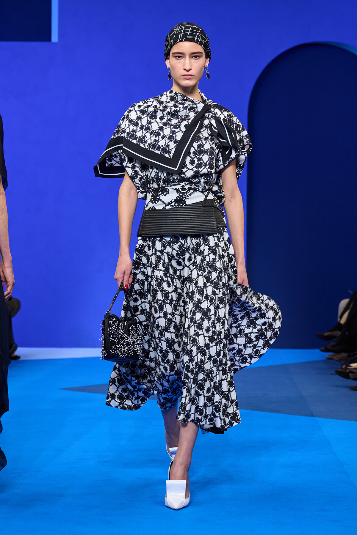 Model on a blue runway wearing a black-and-white patterned dress with a matching headscarf and bold belt, carrying a small studded bag and white heels.